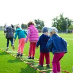 Children enjoy an outdoor activity on a grassy field, stepping over a ladder.