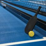 Pickleball paddle resting against net with yellow ball on blue court